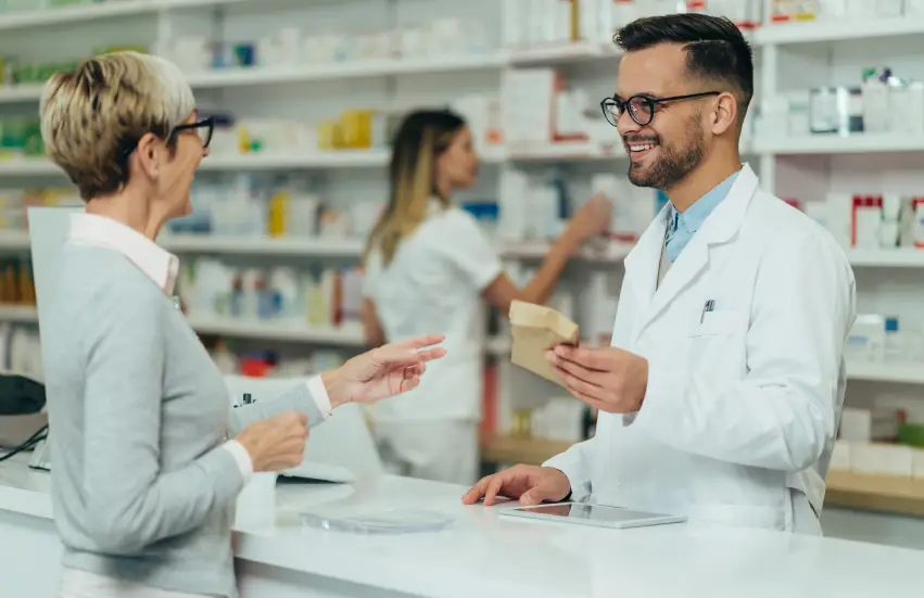 Young male pharmacist giving prescription medications to senior female customer in a pharmacy with female pharmacist in the background